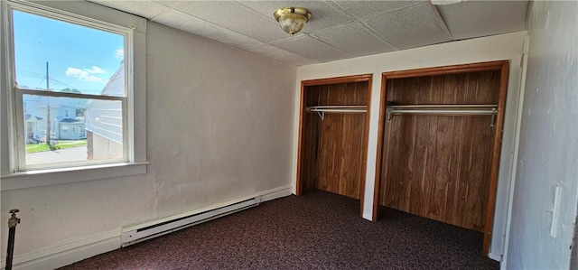 unfurnished bedroom with dark colored carpet, a drop ceiling, two closets, and baseboard heating