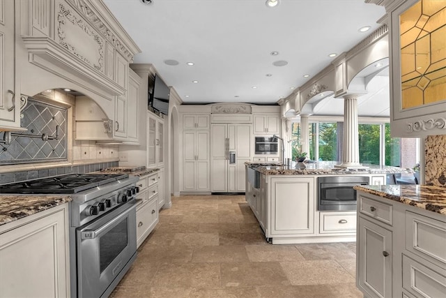 kitchen with dark stone counters, decorative columns, built in appliances, light tile flooring, and backsplash