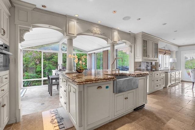 kitchen with vaulted ceiling, a center island with sink, light stone countertops, ornate columns, and light tile floors