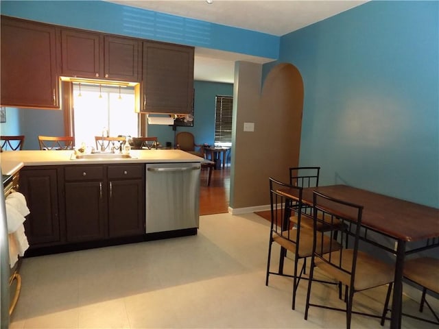 kitchen featuring arched walkways, light countertops, stainless steel dishwasher, a sink, and dark brown cabinets