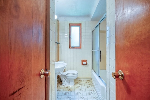 bathroom featuring toilet, bath / shower combo with glass door, and tile walls