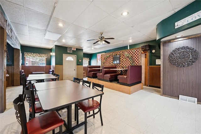 dining space with a drop ceiling, wooden walls, and ceiling fan