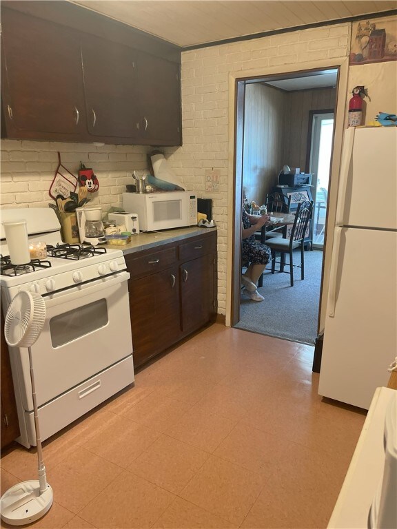 kitchen with light carpet, dark brown cabinetry, white appliances, and brick wall