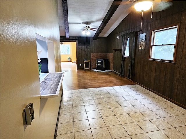 interior space featuring ceiling fan, beamed ceiling, a wood stove, light tile floors, and wood walls