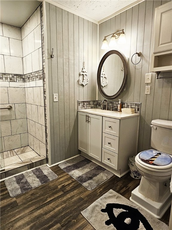 bathroom with a tile shower, hardwood / wood-style floors, wood walls, vanity with extensive cabinet space, and toilet