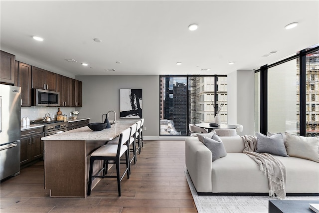 kitchen with a kitchen breakfast bar, a center island with sink, a wall of windows, dark wood-type flooring, and appliances with stainless steel finishes