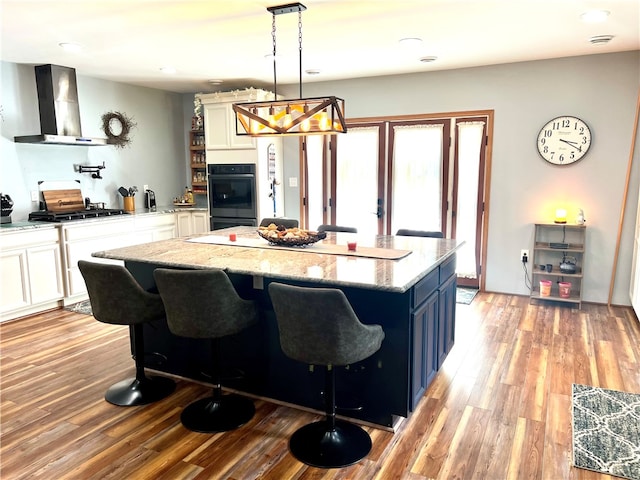 kitchen with pendant lighting, wall chimney range hood, blue cabinetry, appliances with stainless steel finishes, and a kitchen bar