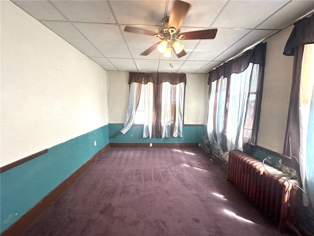 unfurnished room with carpet flooring, ceiling fan, radiator heating unit, and a drop ceiling