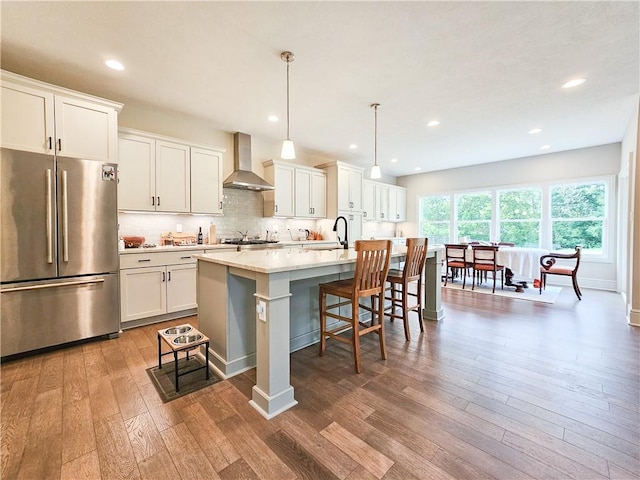 kitchen featuring a center island with sink, hanging light fixtures, wall chimney exhaust hood, white cabinetry, and stainless steel refrigerator