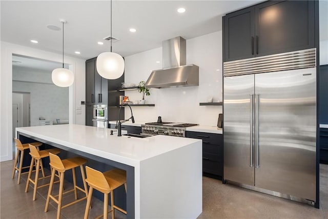 kitchen with appliances with stainless steel finishes, sink, wall chimney range hood, pendant lighting, and a center island with sink