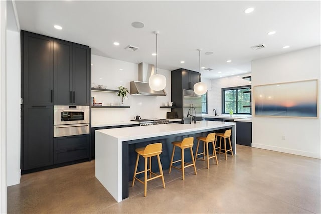 kitchen with stainless steel appliances, wall chimney range hood, pendant lighting, a breakfast bar area, and a center island with sink