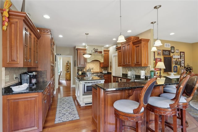 kitchen with a kitchen breakfast bar, light hardwood / wood-style floors, dark stone countertops, backsplash, and hanging light fixtures