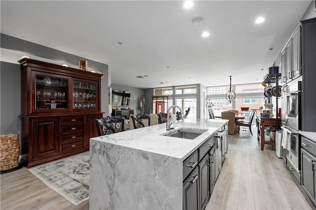 kitchen with light stone counters, a sink, gray cabinets, light wood finished floors, and an island with sink