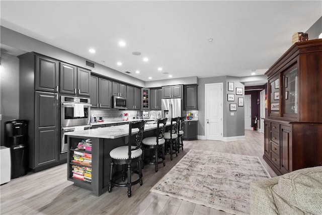 kitchen featuring a breakfast bar area, appliances with stainless steel finishes, backsplash, light wood finished floors, and an island with sink