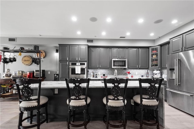 kitchen featuring a center island with sink, appliances with stainless steel finishes, a breakfast bar area, light stone countertops, and gray cabinetry