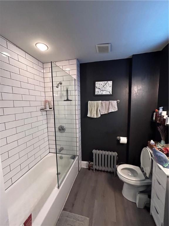 full bathroom with radiator heating unit, bath / shower combo with glass door, wood-type flooring, toilet, and vanity