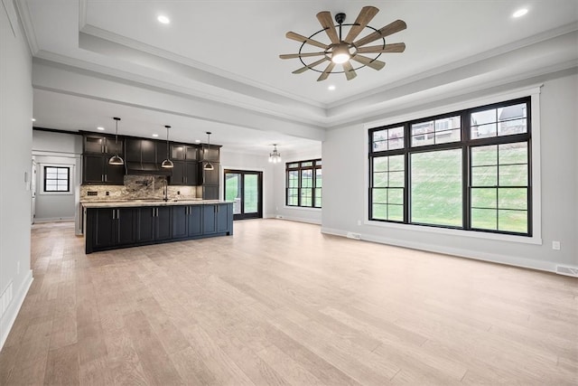 unfurnished living room with ceiling fan with notable chandelier, a raised ceiling, sink, crown molding, and light hardwood / wood-style flooring