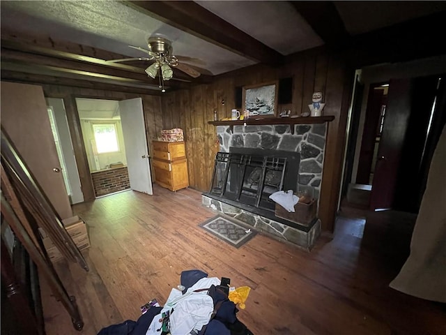 living room with ceiling fan, wooden walls, hardwood / wood-style flooring, beamed ceiling, and a stone fireplace