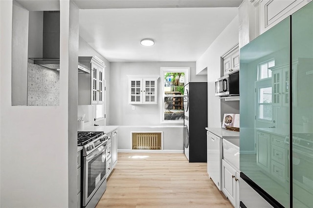 kitchen with stainless steel appliances, wall chimney range hood, white cabinets, and light hardwood / wood-style flooring