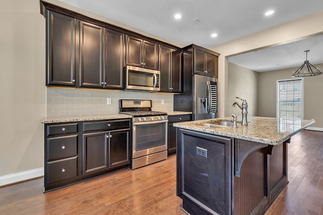 kitchen featuring tasteful backsplash, hardwood / wood-style flooring, stainless steel appliances, light stone counters, and a kitchen island with sink