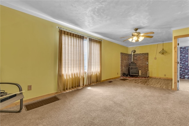 unfurnished living room with a wood stove, carpet floors, crown molding, and ceiling fan