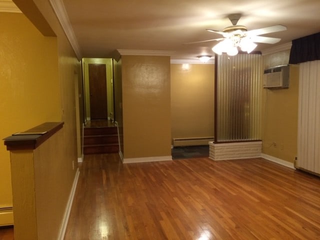 spare room with a wall unit AC, hardwood / wood-style floors, baseboard heating, ceiling fan, and ornamental molding