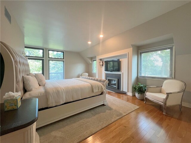 bedroom with light wood-type flooring, vaulted ceiling, multiple windows, and a glass covered fireplace