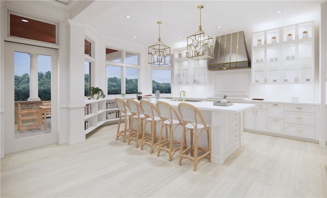 kitchen featuring white cabinetry, wall chimney range hood, a kitchen breakfast bar, decorative light fixtures, and a center island with sink