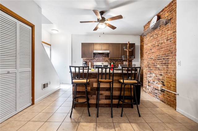 interior space featuring light tile patterned floors, ceiling fan, brick wall, decorative backsplash, and dark brown cabinetry