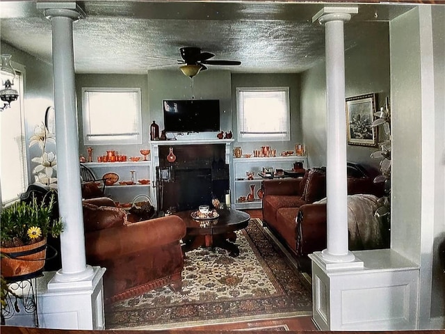 living area featuring ceiling fan, a fireplace, ornate columns, and a textured ceiling