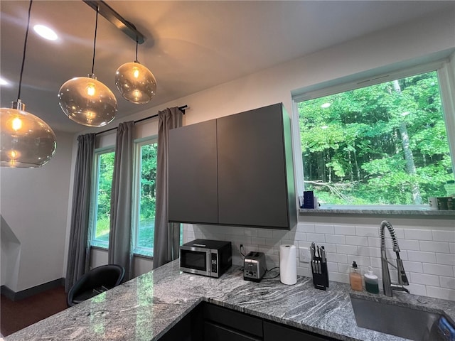 kitchen featuring stone counters, a sink, backsplash, stainless steel microwave, and decorative light fixtures