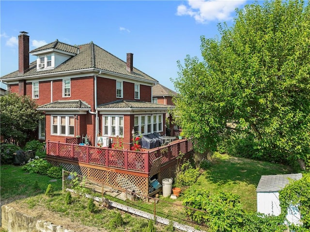 rear view of house featuring a yard, a wooden deck, brick siding, a chimney, and a tiled roof