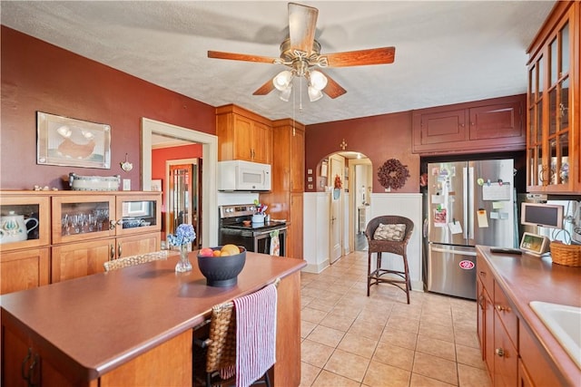 kitchen with light tile patterned floors, a ceiling fan, arched walkways, glass insert cabinets, and appliances with stainless steel finishes