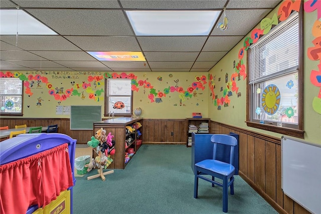 recreation room with wooden walls, a drop ceiling, carpet flooring, and wainscoting