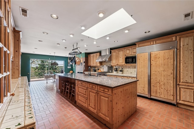 kitchen with decorative backsplash, built in appliances, wall chimney range hood, a skylight, and an island with sink