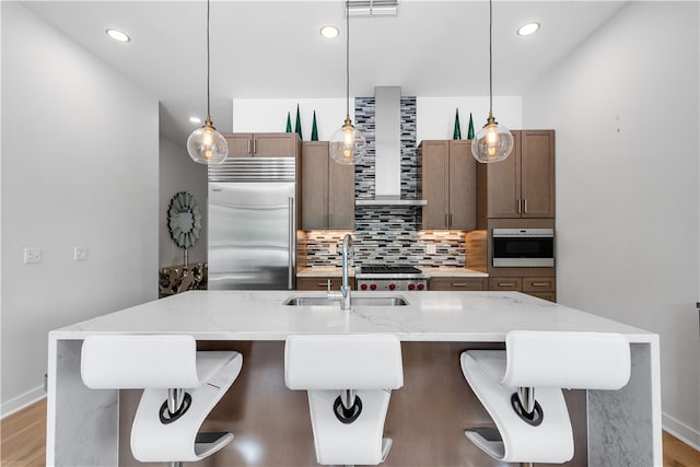 kitchen with decorative light fixtures, light stone counters, stainless steel appliances, wall chimney range hood, and a center island with sink