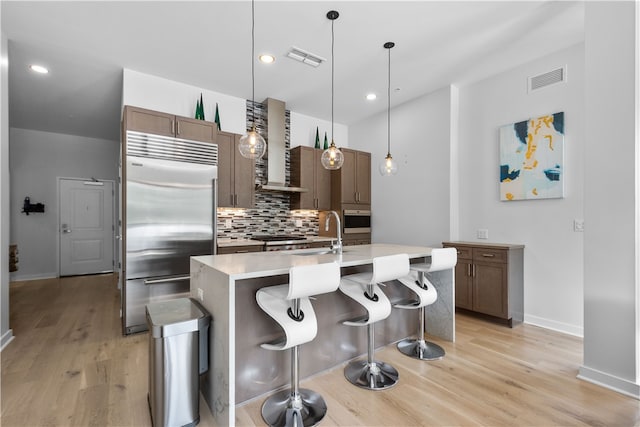 kitchen featuring wall chimney exhaust hood, pendant lighting, light hardwood / wood-style floors, a kitchen island with sink, and stainless steel appliances