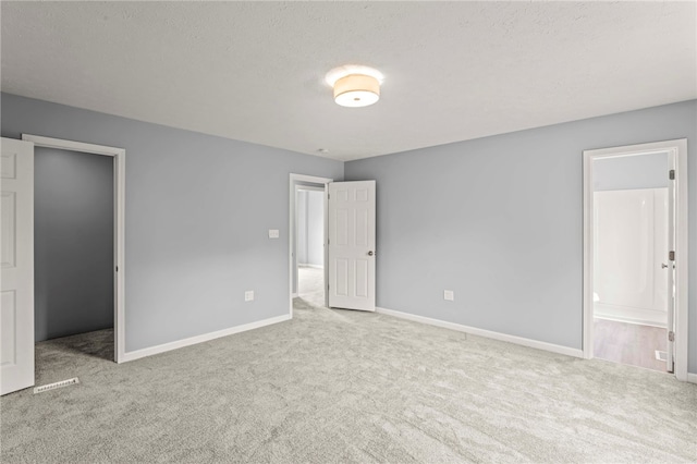 unfurnished bedroom featuring light colored carpet, a spacious closet, a textured ceiling, ensuite bath, and baseboards