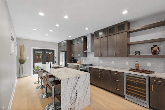 kitchen with beverage cooler, sink, a kitchen island with sink, wall chimney exhaust hood, and light wood-type flooring