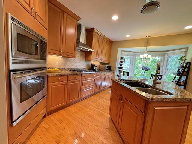 kitchen with light wood-type flooring, wall chimney exhaust hood, stainless steel appliances, sink, and an island with sink