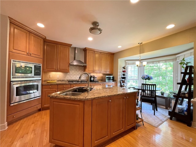 kitchen featuring wall chimney range hood, light hardwood / wood-style flooring, light stone countertops, an island with sink, and decorative light fixtures