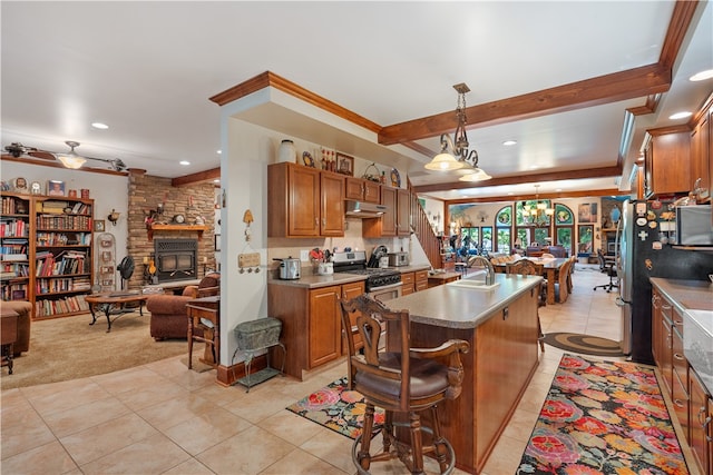 kitchen featuring light colored carpet, a stone fireplace, an island with sink, decorative light fixtures, and stainless steel gas stove