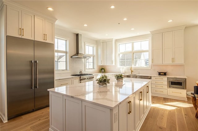 kitchen with stainless steel built in fridge, light wood finished floors, wall chimney exhaust hood, and light stone countertops