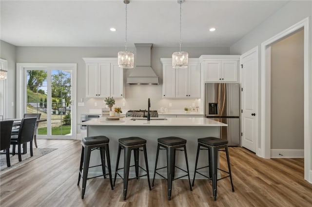 kitchen with light hardwood / wood-style floors, a kitchen island with sink, premium range hood, stainless steel fridge with ice dispenser, and white cabinets