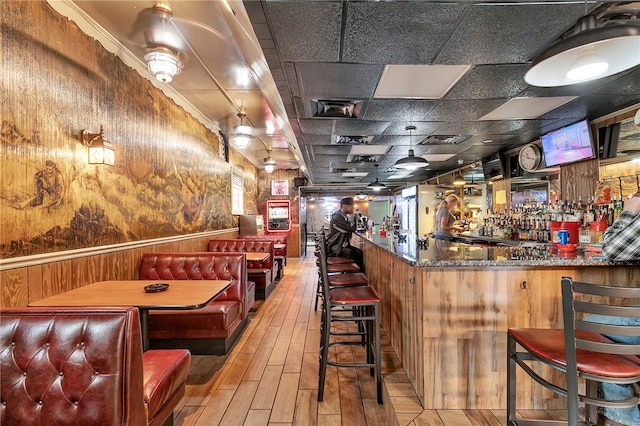 bar featuring light wood-type flooring, dark stone counters, a paneled ceiling, and wooden walls