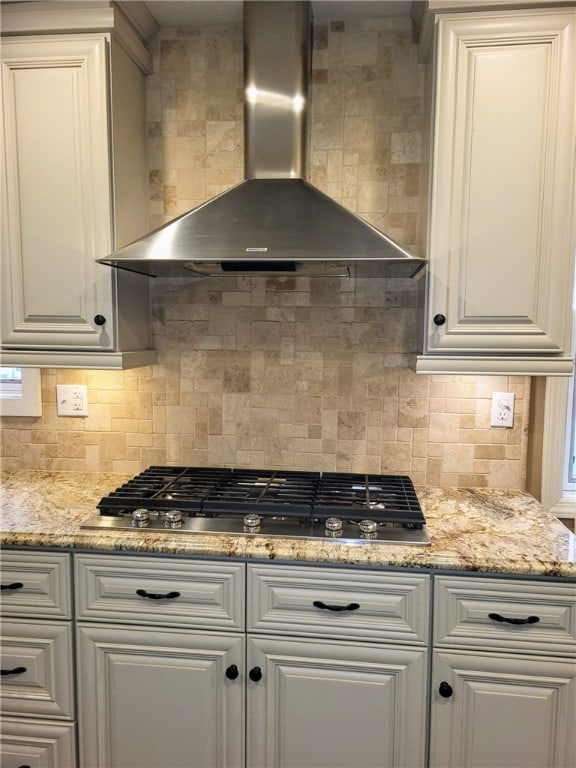 kitchen featuring wall chimney range hood, decorative backsplash, stainless steel gas stovetop, white cabinetry, and light stone countertops