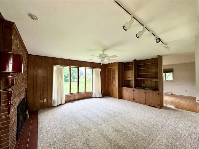 unfurnished living room with ceiling fan, wood walls, a brick fireplace, rail lighting, and carpet floors