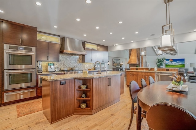 kitchen featuring light hardwood / wood-style floors, stainless steel appliances, wall chimney exhaust hood, and an island with sink