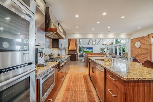 kitchen with appliances with stainless steel finishes, a large island with sink, light hardwood / wood-style floors, wall chimney exhaust hood, and tasteful backsplash