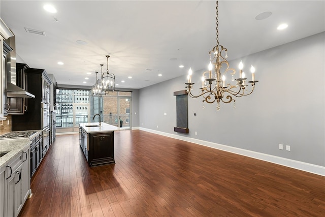 kitchen featuring dark wood-type flooring, a center island with sink, wall chimney range hood, decorative light fixtures, and stainless steel appliances